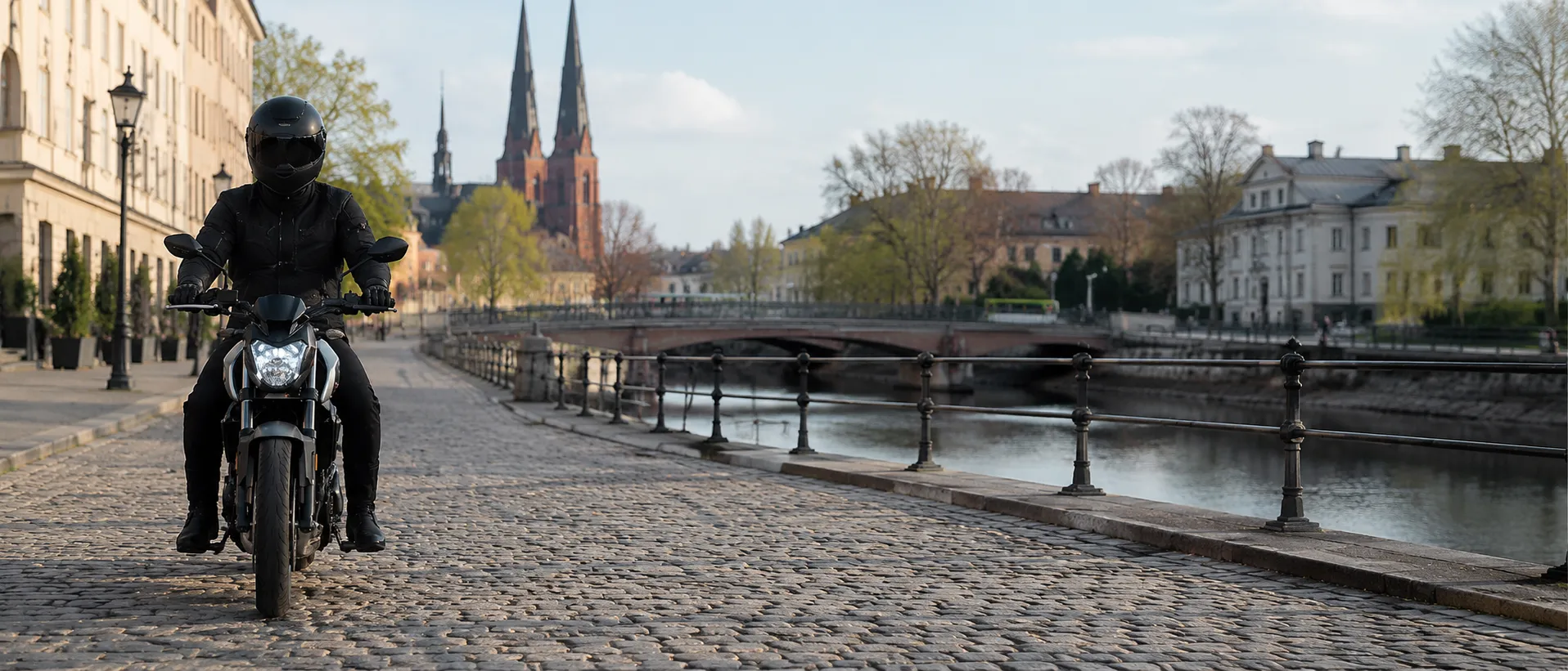 Förare på lätt motorcykel vid Fyrisån i Uppsala med domkyrkan i bakgrunden.
